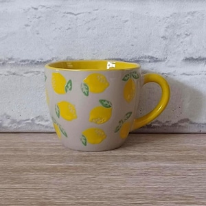 Pode incluir: Uma caneca de cerâmica com exterior cinza claro e interior e alça amarelo brilhante. A caneca é decorada com limões amarelos e folhas verdes. A caneca está sobre uma superfície de madeira.