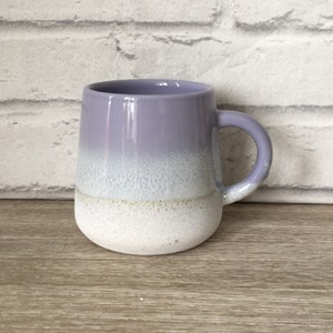 May include: A light purple and white ombre ceramic mug with a textured finish. The mug has a rounded shape and a handle.