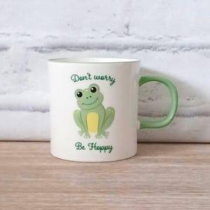 May include: White ceramic mug with a green handle and rim. The mug features a cartoon frog illustration with the text "Don't worry, Be Hoppy" in green script. The frog is light green with a yellow belly.
