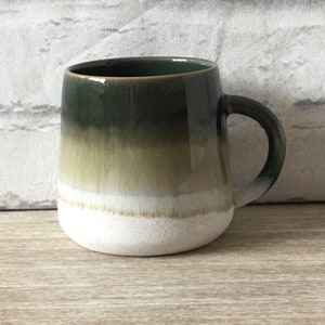 Puede incluir: Una taza de cerámica con forma redondeada y asa. La taza presenta un degradado de colores, comenzando con un verde oscuro en la parte superior, pasando a un verde oliva claro y terminando con una base blanca. La taza está diseñada para bebidas calientes.