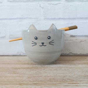 May include: A light gray ceramic bowl shaped like a cat's face, with ears, eyes, nose, and whiskers. A pair of wooden chopsticks rests across the top of the bowl. The bowl is on a wooden surface.