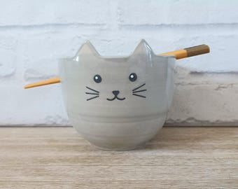 May include: A light gray ceramic bowl shaped like a cat's face, with ears, eyes, nose, and whiskers. A pair of wooden chopsticks rests across the top of the bowl. The bowl is on a wooden surface.