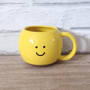 May include: A yellow ceramic mug with a smiling face design. The mug has a rounded shape, a brown base, and a curved handle. The background is a white brick wall and a wooden surface.