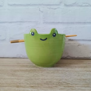May include: A green ceramic bowl shaped like a frog with a smiling face and two small, round eyes. A pair of chopsticks is placed across the bowl. The bowl is on a wooden surface.