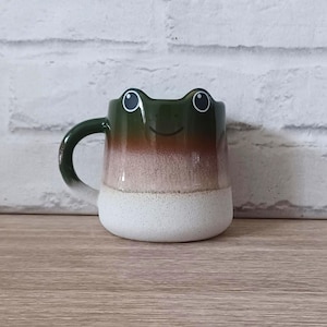 May include: A ceramic frog-shaped mug with a green top, brown mid-section, and white base. The frog's face has large, round eyes and a smiling mouth. The handle is green.