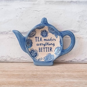 Puede incluir: Un posa-bolsas de té de cerámica azul con forma de tetera, con diseños florales y el texto "TEA makes everything BETTER". El soporte tiene un asa y un pico, sobre un fondo de ladrillos blancos.