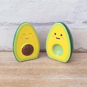 May include: Two avocado-shaped ceramic figurines with smiling faces. The avocados have a green exterior, yellow interior, and a brown pit. One has a pit, the other has a hole. The figurines are on a wooden surface.