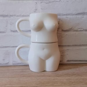 May include: Two white ceramic mugs stacked together, shaped like a female torso. The top mug depicts the upper body with handles, while the bottom mug forms the lower body. The mugs are smooth and glossy, with a minimalist design.