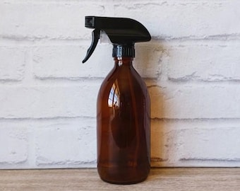 May include: Amber glass spray bottle with a black trigger sprayer. The bottle has a rounded shape and is sitting on a wooden surface. The background is a white brick wall.