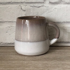 Puede incluir: Una taza de cerámica con un esmalte degradado blanco y marrón. La taza tiene una forma redondeada y un asa.