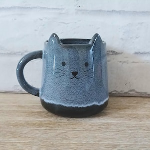 Op de afbeelding: Een grijze keramische mok in de vorm van een kat, met oren aan de bovenkant en een kattengezicht. De mok heeft een donkergrijze basis en handvat en staat op een houten oppervlak.