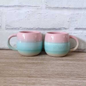 May include: Two ceramic mugs with a rounded shape and a handle. Each mug features three horizontal colour bands: pink at the top, light blue in the middle, and a cream colour at the bottom. The mugs are placed on a wooden surface.