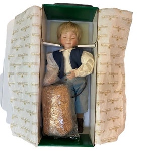 May include: A porcelain doll of a young boy with blonde hair, wearing a white shirt, blue vest, and blue pants. The doll is in a box with printed tissue paper. A bundle of straw is wrapped in plastic.