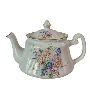 Puede incluir: Tetera blanca con un diseño floral con flores rosas, azules y amarillas. La tetera tiene un borde dorado alrededor de la tapa y el pico.