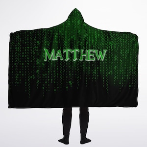 May include: A black hooded blanket featuring a green digital rain design and the name "MATTHEW" in green. The blanket has a hood and is worn by a person. The design is reminiscent of computer code.