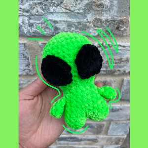 May include: A green crocheted alien plush toy with black eyes. The toy is being held in a hand.