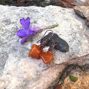 Puede incluir: Un par de pendientes con piedras de color naranja en forma de lágrima y detalles de alambre de cobre. Los pendientes están sobre una roca con una flor morada.