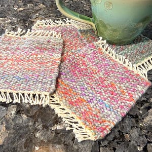 May include: A light green ceramic mug rests on three colorful woven coasters. The coasters are rectangular with a fringed edge and a speckled pattern of pink, orange, and blue threads. The mug has a curved handle.