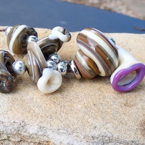 May include: A collection of handmade glass beads in various shapes and colors. The beads feature swirls of brown, white, and tan, with some having a purple accent. Silver-colored metal beads and a brown cord are also present.