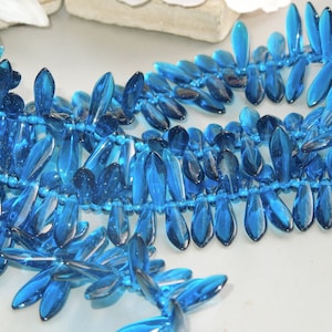 May include: A strand of teal blue glass beads in a leaf shape. The beads are strung on a clear thread and are arranged in a staggered pattern.
