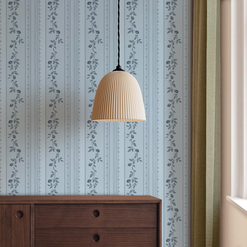May include: A cream-coloured, ribbed pendant lamp suspended from a black cord against light blue floral wallpaper. A dark brown wooden cabinet is below the wallpaper. Green curtains are on the right.