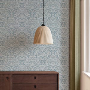 May include: A cream-coloured, ribbed pendant light hangs from a black cord against a light blue wallpaper with a white floral pattern. A dark brown wooden dresser is visible below, and a green curtain is on the right.