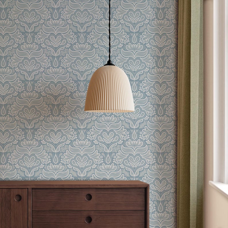 May include: A cream-coloured, ribbed pendant light hangs from a black cord against a light blue wallpaper with a white floral pattern. A dark brown wooden dresser is visible below, and a green curtain is on the right.