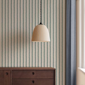 May include: A cream-coloured, ribbed pendant light hangs from a black cord. The light is centred in front of a wall with vertical stripes of pale green on a cream background. A dark brown wooden cabinet is below the light, and a blue curtain is to the right.