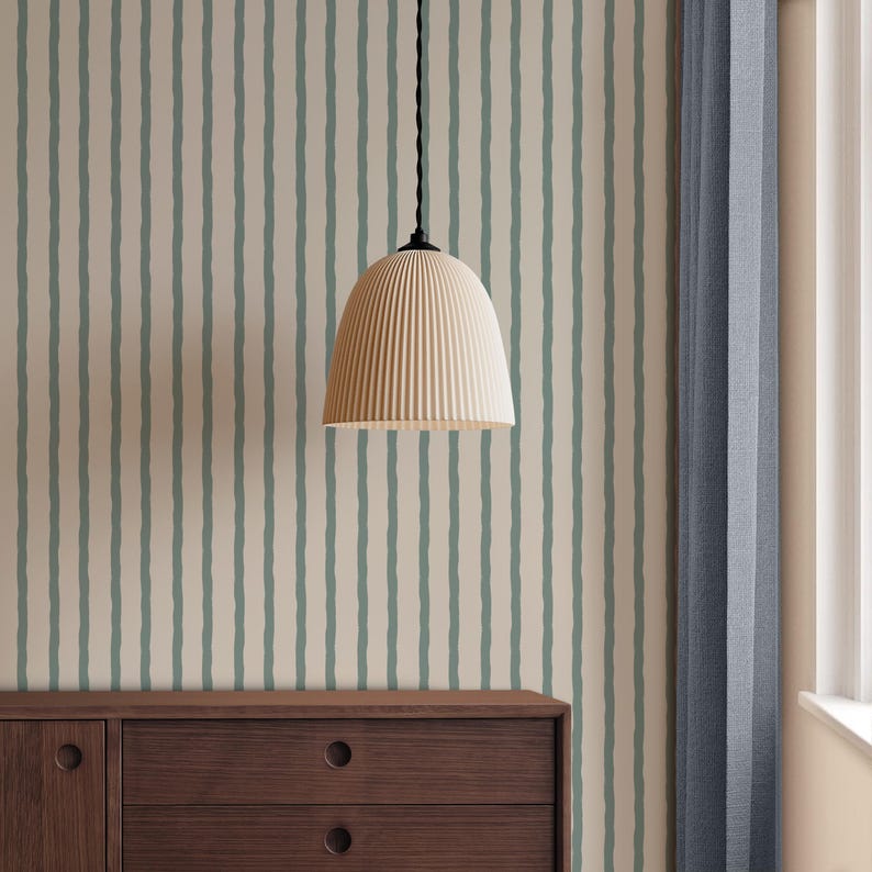 May include: A cream-coloured, ribbed pendant light hangs from a black cord. The light is centred in front of a wall with vertical stripes of pale green on a cream background. A dark brown wooden cabinet is below the light, and a blue curtain is to the right.