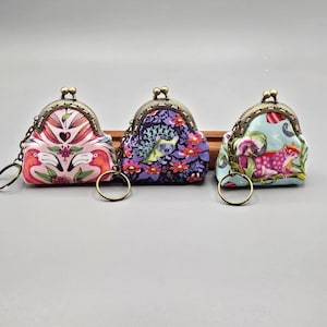 May include: Three small, patterned coin purses with kiss-lock closures and key rings. The purses feature colorful floral and animal designs, including flamingos, a deer, and a pink spotted animal. The metal frames and chains have a bronze finish.