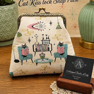 May include: A cream-colored fabric kiss lock snap purse featuring a printed pattern of two black cats reading books in teal armchairs, a bookshelf, and starburst motifs. The purse has an antique bronze-toned metal frame and a top snap closure.