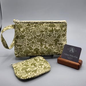 May include: A set of two olive green fabric pouches with a mushroom and leaf pattern. One pouch is a larger zippered bag with a handle, and the other is a smaller coin purse. A business card with the text "Antiqued Basket Lady" is in a wooden holder.