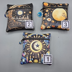 May include: Three small, square, black zippered pouches with celestial designs. One features a crescent moon and star, another a full moon and flowers, and the third a night sky scene. Each has a star-shaped zipper pull and a numbered tag.