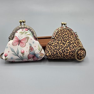 May include: Two small coin purses with kiss-lock closures. One purse features a white background with pink and blue butterflies and floral accents. The other purse has a brown and tan leopard print pattern. Both purses have metal frames and key rings.