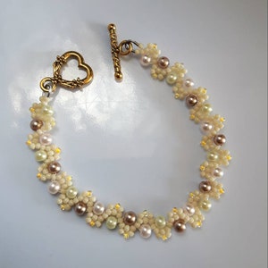 May include: A delicate bracelet made with small white and brown beads, forming a floral pattern. The bracelet has a heart-shaped clasp and a toggle closure.