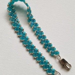May include: A bracelet crafted from translucent turquoise square beads and small metallic silver seed beads arranged in a repeating woven pattern. The piece features a silver-toned lobster claw clasp at one end and a small metal ring at the other.