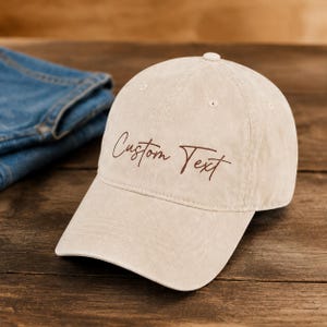 Op de afbeelding: Beige baseballpet met de tekst "Custom Text" in bruin script. De pet ligt op een houten oppervlak naast een spijkerbroek.