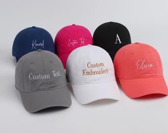 May include: Six baseball caps in a range of colours: navy, pink, black, grey, white, and coral. Each cap has custom embroidery with names or text. The caps are displayed on a white surface.