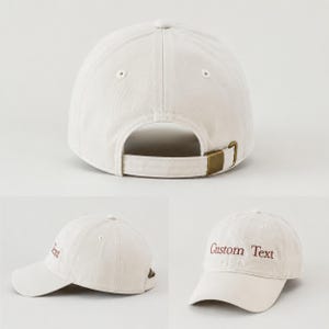 May include: Off-white baseball cap with a curved brim and adjustable strap. The cap features the embroidered text "Custom Text" in a dark red color. The cap has a metal buckle closure.