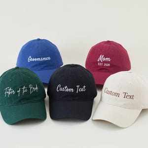 Könnte beinhalten: Fünf Baseballkappen in verschiedenen Farben: Blau, Grün, Schwarz, Burgunderrot und Off-White. Jede Kappe hat einen gestickten Text, darunter "Groomsmen", "Father of the Bride", "Custom Text" und "Mom EST 2026". Die Kappen liegen auf einer weißen Oberfläche.