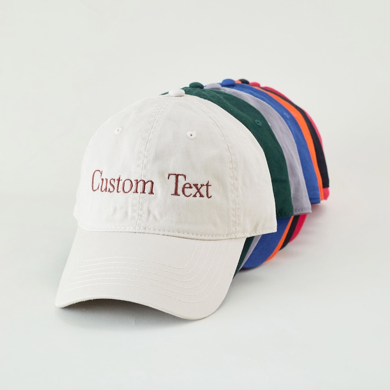 May include: A row of baseball caps in various colors, including white, green, blue, and red. The front cap is off-white with the embroidered text "Custom Text" in a dark red color. The caps are arranged in a diagonal line.