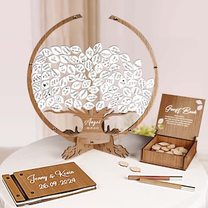 Könnte beinhalten: Ein hölzerner Gästebuchbaum mit weißen Blättern, auf denen die Gäste Nachrichten hinterlassen können. Der Baum steht auf einem weißen Tisch mit einem hölzernen Gästebuch, einer Schachtel mit zusätzlichen Blättern und zwei Stiften. Das Gästebuch ist mit den Namen "Jenny & Kevin" und dem Datum "26.09.2024" graviert.