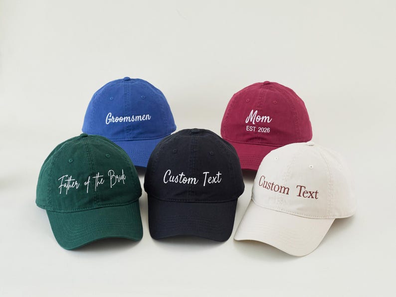 May include: Five baseball caps in various colors: blue, green, black, burgundy, and off-white. Each cap has embroidered text, including "Groomsmen," "Father of the Bride," "Custom Text," and "Mom EST 2026." The caps are arranged on a white surface.