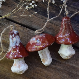 May include: Three ceramic mushroom ornaments with brown and white speckled glaze. The mushrooms are hanging from twine and are displayed on a wooden surface.