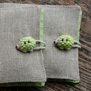 May include: Two beige linen pouches with green trim and white floral designs. Each pouch has a green and white ceramic hedgehog button.