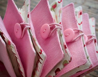 May include: A stack of pink fabric pouches with white fabric lining and a floral pattern. Each pouch has a pink button and a white string closure.
