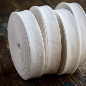 May include: Four spools of white cotton twill tape, rolled up and stacked on top of each other. The tape is a plain weave fabric with a subtle herringbone pattern.