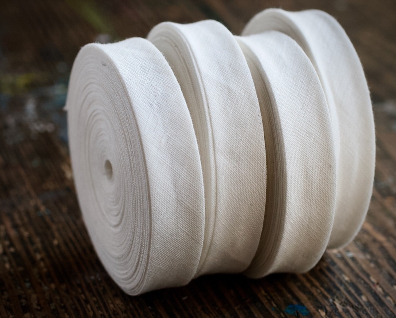 May include: Four spools of white cotton twill tape, rolled up and stacked on top of each other. The tape is a plain weave fabric with a subtle herringbone pattern.