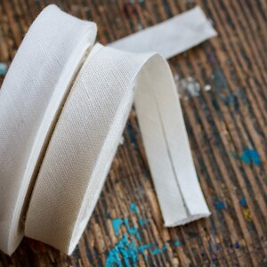 May include: Two rolls of white cotton twill tape, one wider than the other, are lying on a wooden surface. The tape is a woven fabric with a diagonal pattern.