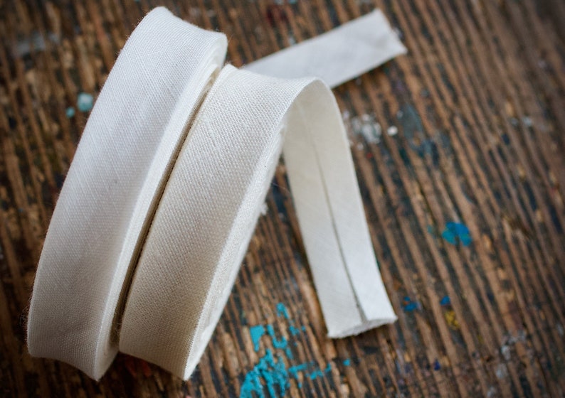 May include: Two rolls of white cotton twill tape, one wider than the other, are lying on a wooden surface. The tape is a woven fabric with a diagonal pattern.